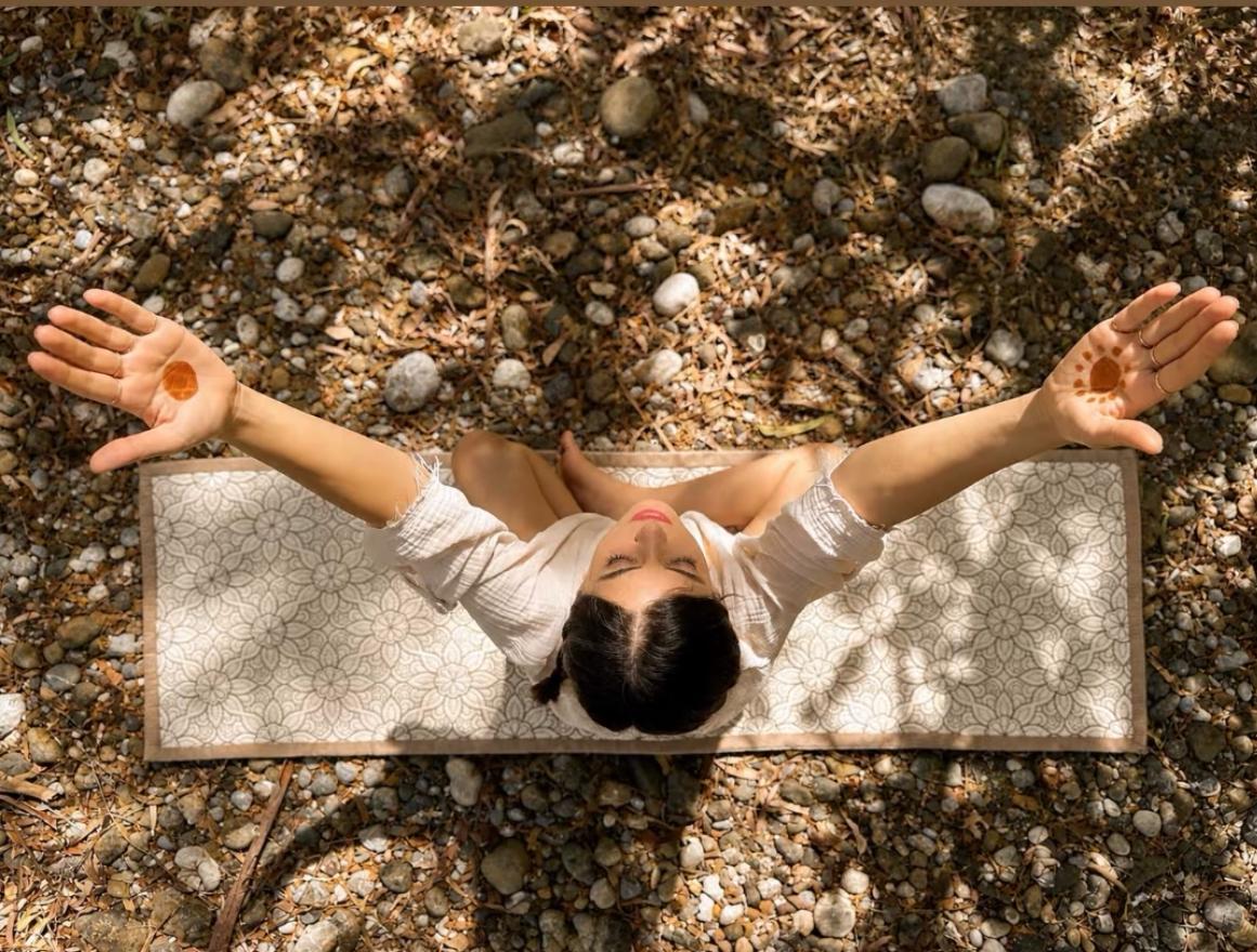 Bovenaanzicht van een vrouw in kleermakerszit met geheven armen op een crèmekleurige Lotus yogamat met bloemenpatroon op kiezelgrond.