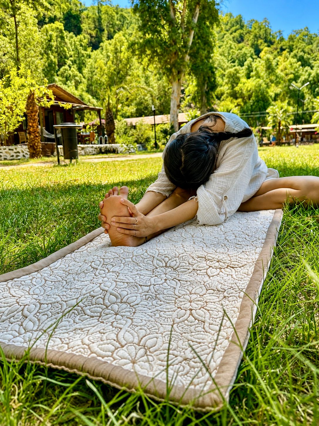 Vrouw die voorover rekt op een crèmekleurige Lotus yogamat op gras, met het bloemenpatroon en de beige rand op de voorgrond.