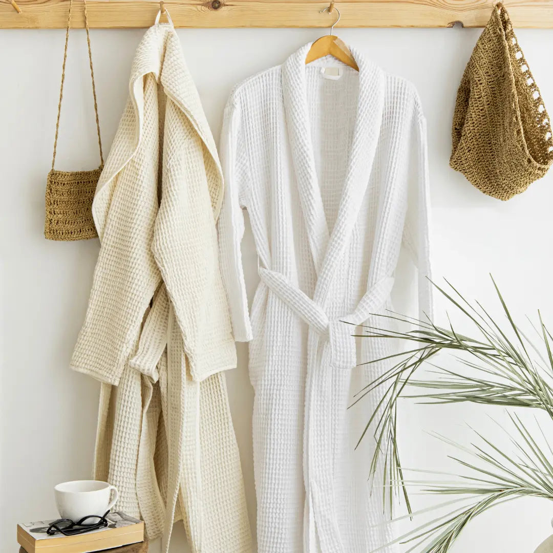 Two Trimita Soft Waffle bathrobes, one beige and one white, hanging on a wooden rack against a white wall with a plant in the foreground.