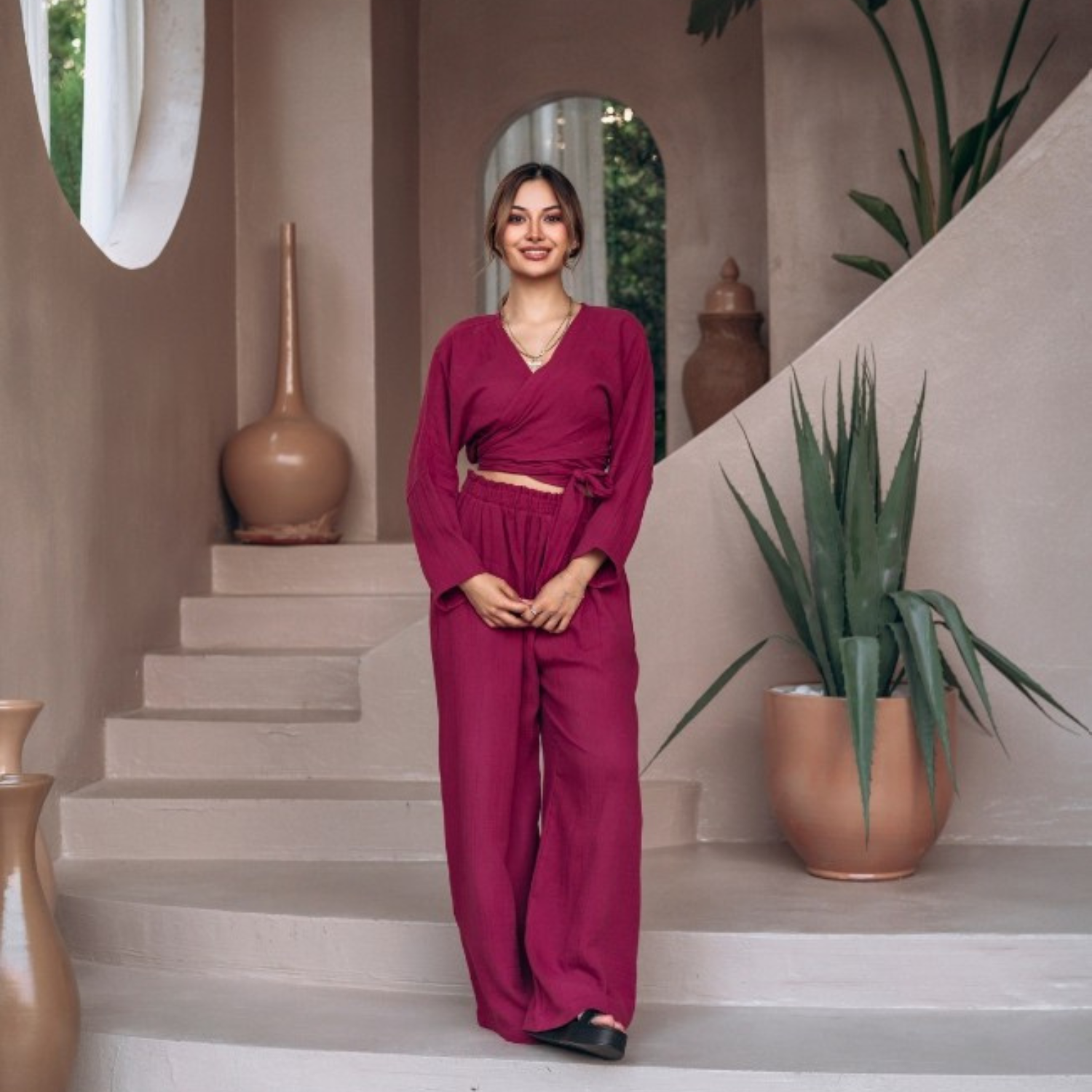 Full-body front view of a smiling woman posing in a vibrant magenta cotton muslin coordinated set, featuring a tie-waist wrap crop top and loose wide-leg trousers in a boho-style resort setting.
