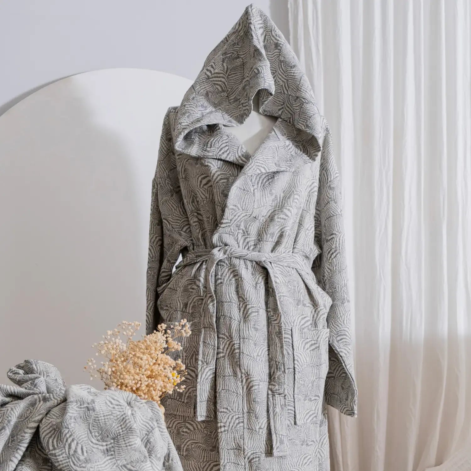A mannequin wearing a light gray muslin hooded bathrobe with a subtle zebra pattern, tied with a belt in a minimalist interior setting.